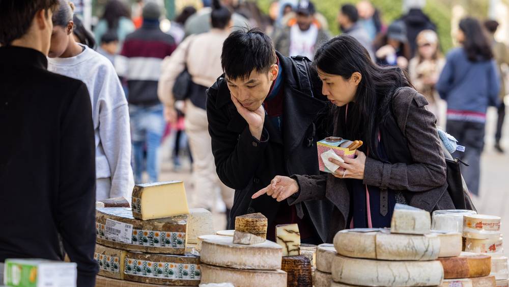 A couple lean in to choose from wheels of Comté and other artisan cheeses at a busy Wembley Park market stall