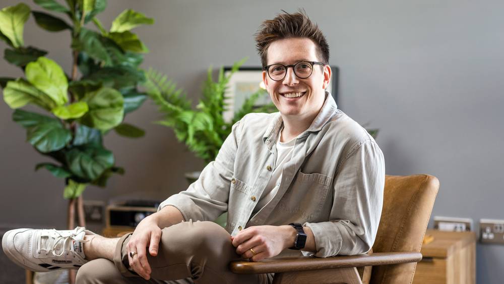 Ben Richards smiling as he relaxes in a tan leather armchair with one leg crossed, beside leafy plants against a grey wall