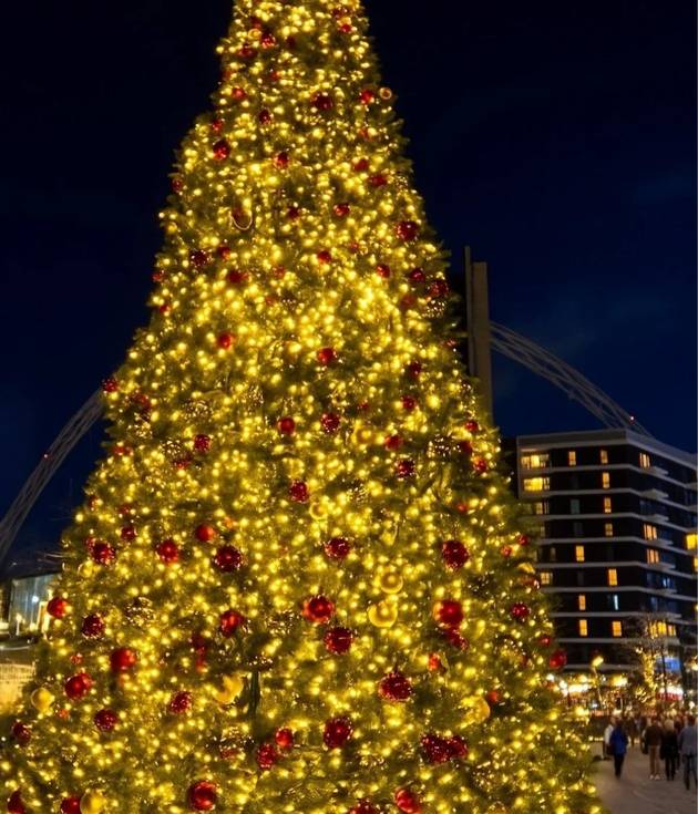 A large Christmas tree covered in golden lights and red baubles in Wembley Park at night, with the Wembley Stadium arch lit blue in the background.