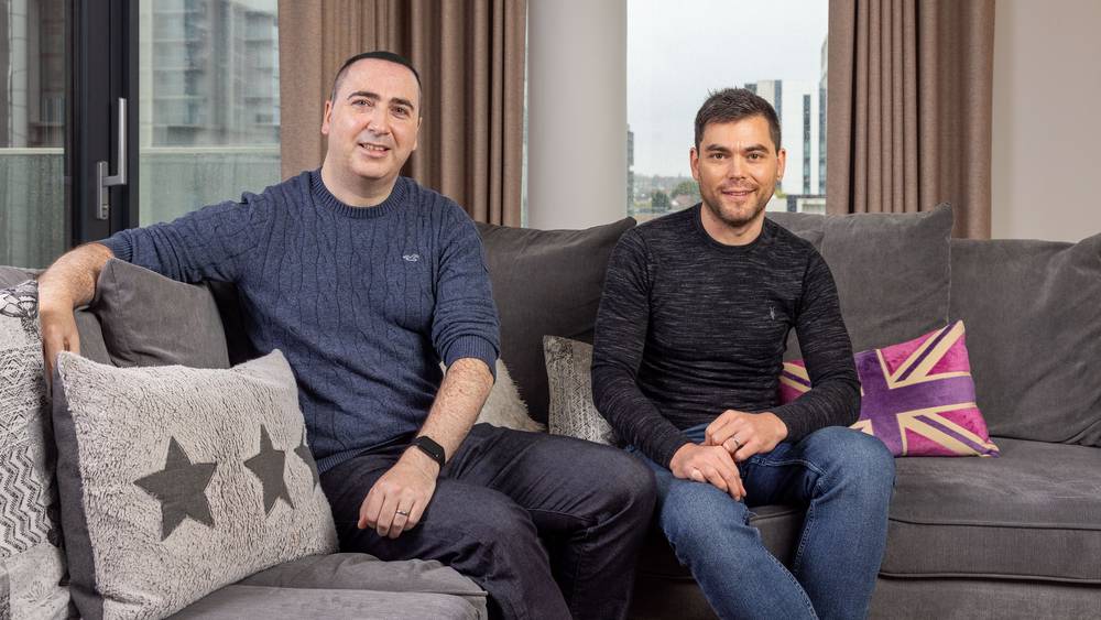 Tim and Conor smiling on a grey corner sofa, one with his arm along the back, beside star and Union Jack cushions, with tall windows behind