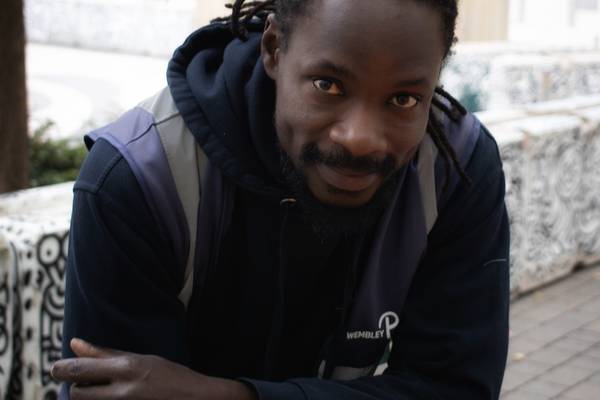 Ebrima Jallow of Wembley Park's cleaning team in a Wembley Park hoodie and hi-vis vest, leaning forward and smiling beside patterned public seating