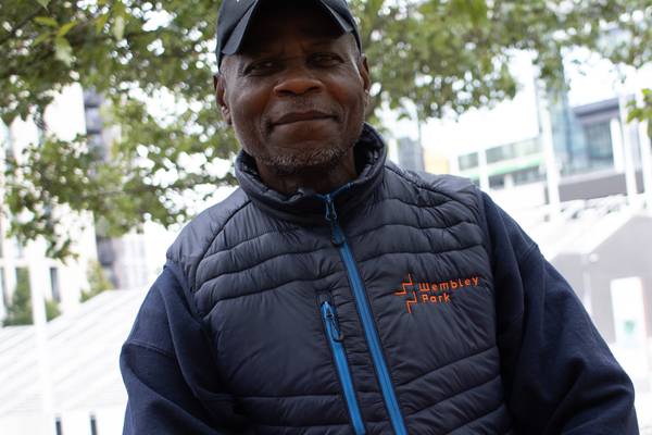 Abdoul Dissou of Wembley Park's cleaning team in a Wembley Park cap and navy gilet, sitting under the trees in Wembley Park